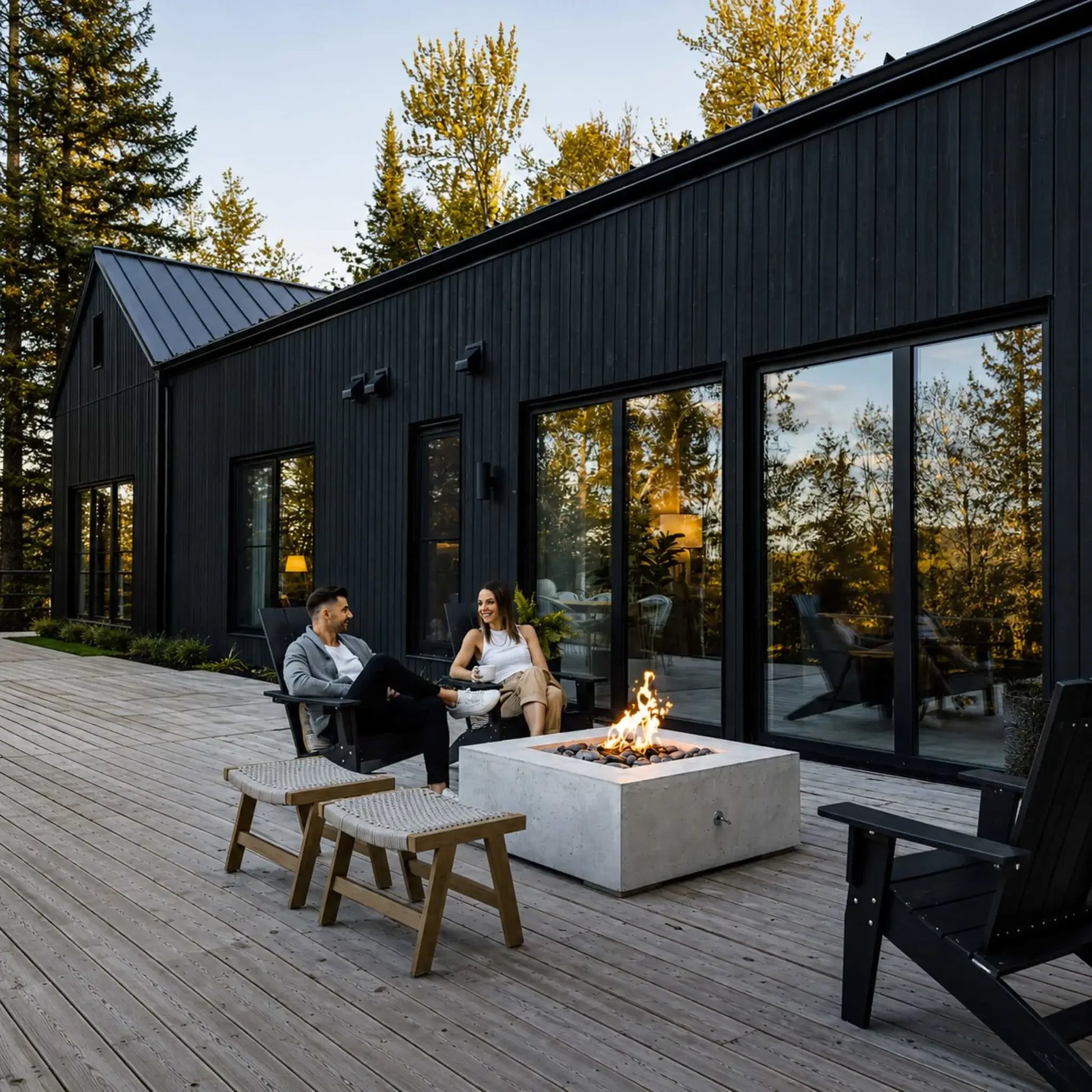 HPC Fire Hearth Products Controls Dekko GFRC Concrete Alea 40 x 40 Fire Pit installed on a spacious wooden deck outside a modern black exterior home. The square concrete fire pit burns with a vibrant flame over decorative stone media while two people relax nearby. Large glass doors, contemporary architecture, and forest views emphasize the upscale outdoor living experience.