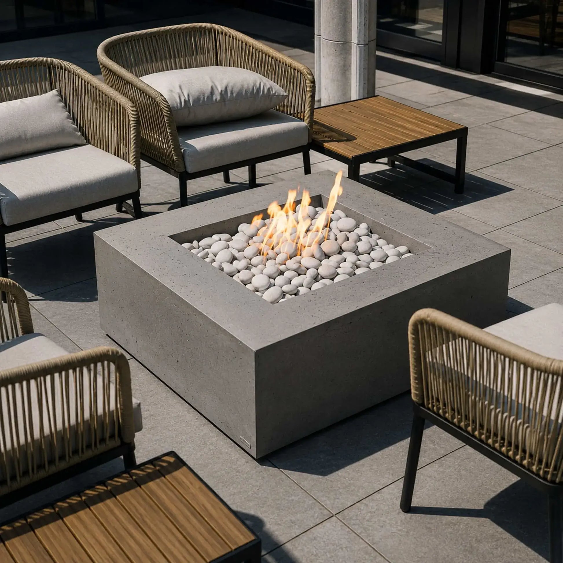 HPC Fire Hearth Products Controls Dekko GFRC Concrete Alea 40 x 40 Fire Pit positioned within a luxury patio furniture arrangement featuring woven lounge chairs and teak side tables. The square GFRC concrete fire pit showcases bright flames rising from smooth white fire stones, creating an elegant centerpiece for sophisticated outdoor seating areas, hospitality spaces, and residential patios.