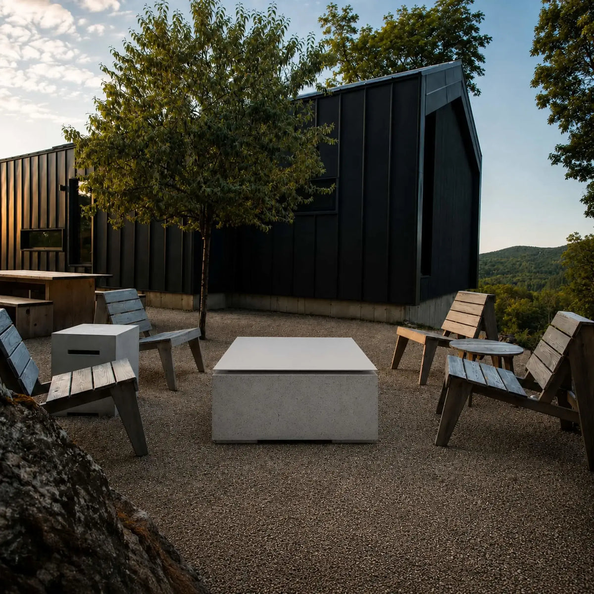 HPC Fire Hearth Products Controls Dekko GFRC Concrete Alea 40 x 40 Fire Pit Table Top Lid showcased in a modern outdoor lifestyle setting. The covered square fire pit sits at the center of a seating area surrounded by wooden lounge chairs on a gravel patio. A contemporary black architectural building, mature trees, and natural landscape create a stylish outdoor gathering space highlighting the versatility of the tabletop lid.