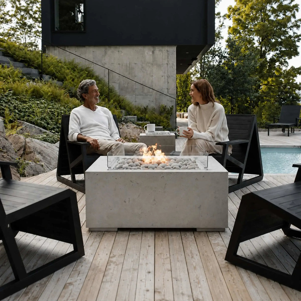 HPC Fire Hearth Products Controls Dekko GFRC Concrete Alea 40 x 40 Fire Pit with tempered glass wind guard displayed in a luxury outdoor living space. A man and woman sit in modern lounge chairs around the lit fire pit, enjoying conversation on a wood deck beside a contemporary home and pool. The image highlights the Alea fire pit’s ivory stone media, clean concrete design, and inviting atmosphere for upscale backyard entertaining.