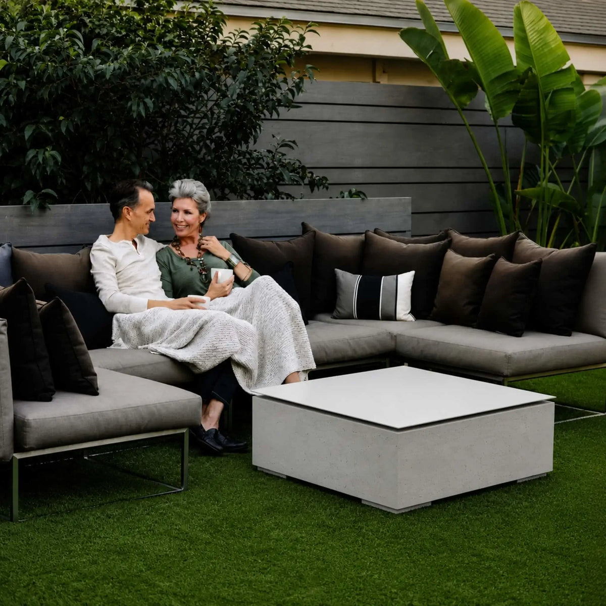 HPC Fire Hearth Products Controls Dekko GFRC Concrete Alea Bravo 32 x 32 Fire Pit Table Top Lid featured in an outdoor lifestyle setting. A modern square fire pit with a light concrete finish and matching tabletop lid serves as the centerpiece of a contemporary patio lounge. Two adults relax on a large sectional sofa surrounded by decorative landscaping, showcasing how the covered fire pit functions as a stylish outdoor coffee table.