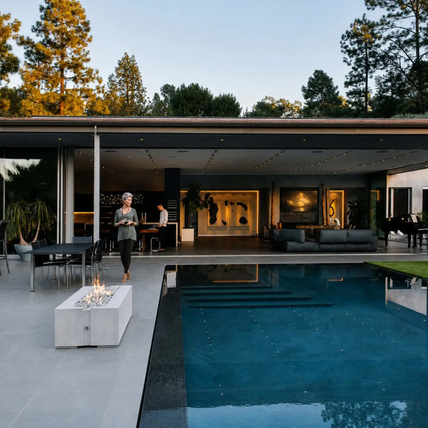 HPC Fire & Hearth Products Controls Dekko GFRC Concrete Avera 48 x 26 Fire Pit featured beside a luxury swimming pool at a modern contemporary residence. The white GFRC concrete fire pit burns with bright flames over decorative stone media, creating a warm focal point along the pool deck. The image showcases the Avera fire pit integrated into an upscale outdoor living environment with expansive architecture, patio seating, and resort-style design.