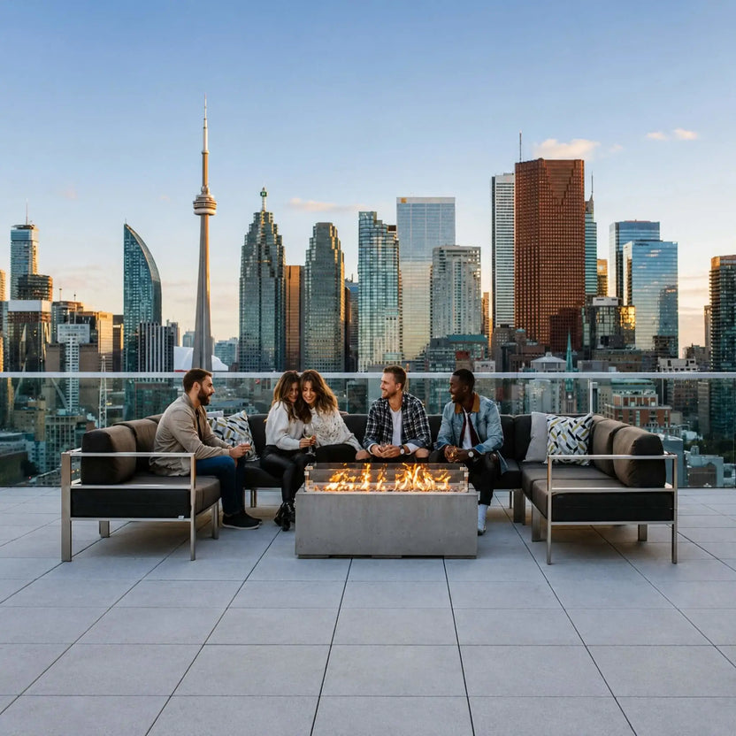 HPC Fire & Hearth Products Controls Dekko GFRC Concrete Avera 48 x 26 Fire Pit serving as the centerpiece of a rooftop gathering space overlooking a city skyline. Several friends sit around the rectangular fire feature enjoying conversation while flames dance across the linear burner. The image highlights the Avera fire pit’s ability to create warmth, atmosphere, and modern outdoor luxury in urban rooftop environments.