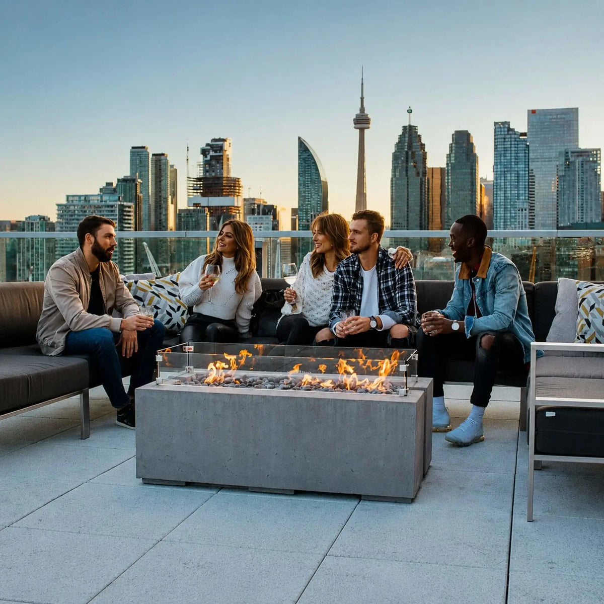HPC Fire & Hearth Products Dekko GFRC Concrete Avera 48 x 26 Fire Pit Tempered Glass Wind Guard Lifestyle 3 image featuring five friends gathered around a modern rectangular concrete fire pit on a rooftop terrace overlooking a dramatic city skyline. The Avera fire pit showcases a tempered glass wind guard, decorative fire media, and vibrant flames, creating a warm social focal point surrounded by contemporary outdoor lounge seating. High-rise buildings and an urban skyline backdrop highlights the fire pit.