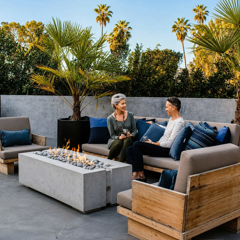Lifestyle image featuring the HPC Fire Hearth Products Controls Dekko GFRC Concrete Avera 60 x 26 Fire Pit in a contemporary outdoor lounge. The rectangular concrete fire pit is filled with decorative stone media and surrounded by cushioned wood-framed seating. Two people relax beside the fire while palm trees and landscaped greenery create an upscale resort-inspired backyard entertainment setting.