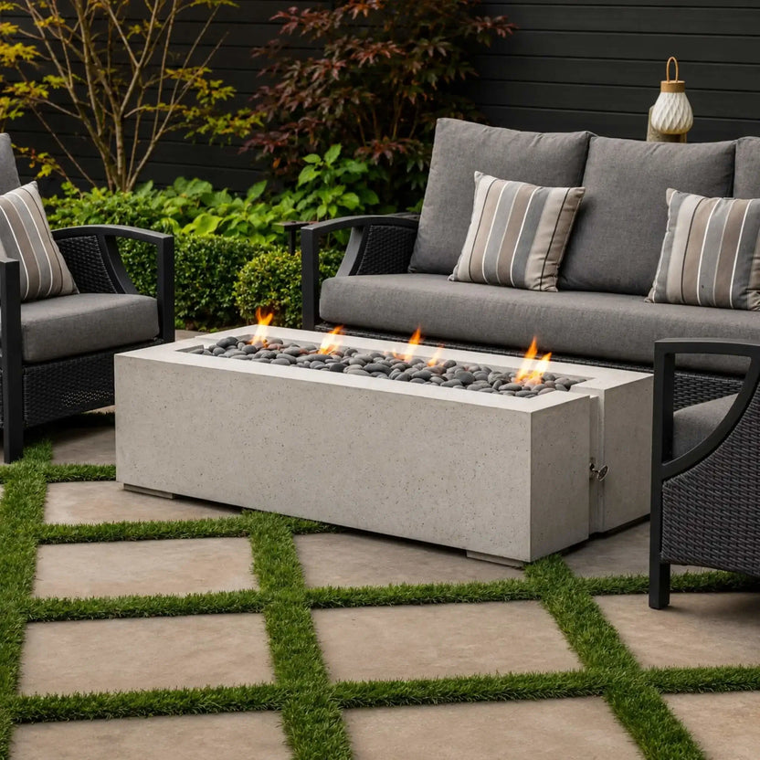 HPC Fire Hearth Products Controls Dekko GFRC Concrete Avera 60 x 26 Fire Pit showcased in a modern patio seating area. The linear concrete fire feature is filled with gray decorative stones and produces a clean flame presentation. Positioned between outdoor lounge chairs and a sofa with neutral cushions, the fire pit complements the landscaped garden and contemporary backyard design.