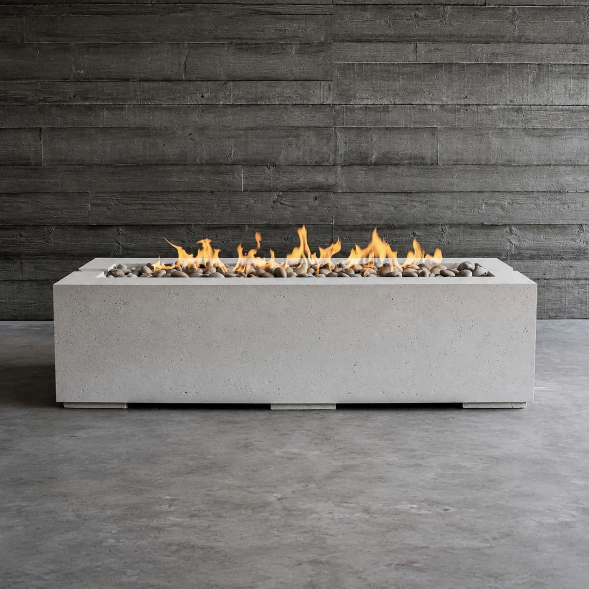 Front-facing lifestyle image of the HPC Fire Hearth Products Controls Dekko GFRC Concrete Avera 60 x 26 Fire Pit burning with a continuous linear flame. Set against a dark wood accent wall, the rectangular concrete fire pit features smooth decorative stones and a minimalist architectural design, making it an ideal centerpiece for modern patios, covered outdoor rooms, and contemporary landscapes.