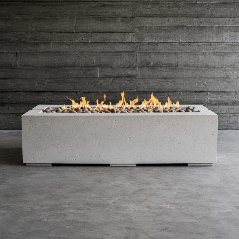 Front-facing lifestyle image of the HPC Fire Hearth Products Controls Dekko GFRC Concrete Avera 60 x 26 Fire Pit burning with a continuous linear flame. Set against a dark wood accent wall, the rectangular concrete fire pit features smooth decorative stones and a minimalist architectural design, making it an ideal centerpiece for modern patios, covered outdoor rooms, and contemporary landscapes.