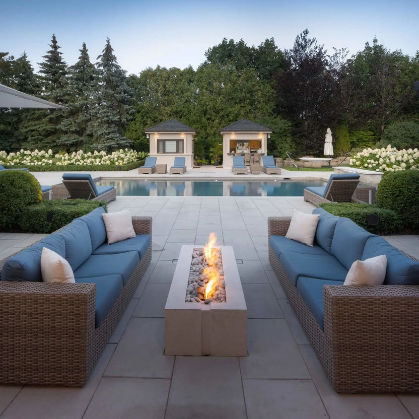 Luxury poolside outdoor living area showcasing the HPC Fire Hearth Products Controls Dekko GFRC Concrete Avera 60 x 26 Fire Pit. Positioned between matching blue-cushioned wicker sofas, the rectangular fire feature provides a focal point overlooking a swimming pool, cabanas, manicured hedges, and landscaped grounds, creating an elegant setting for outdoor entertaining and relaxation.