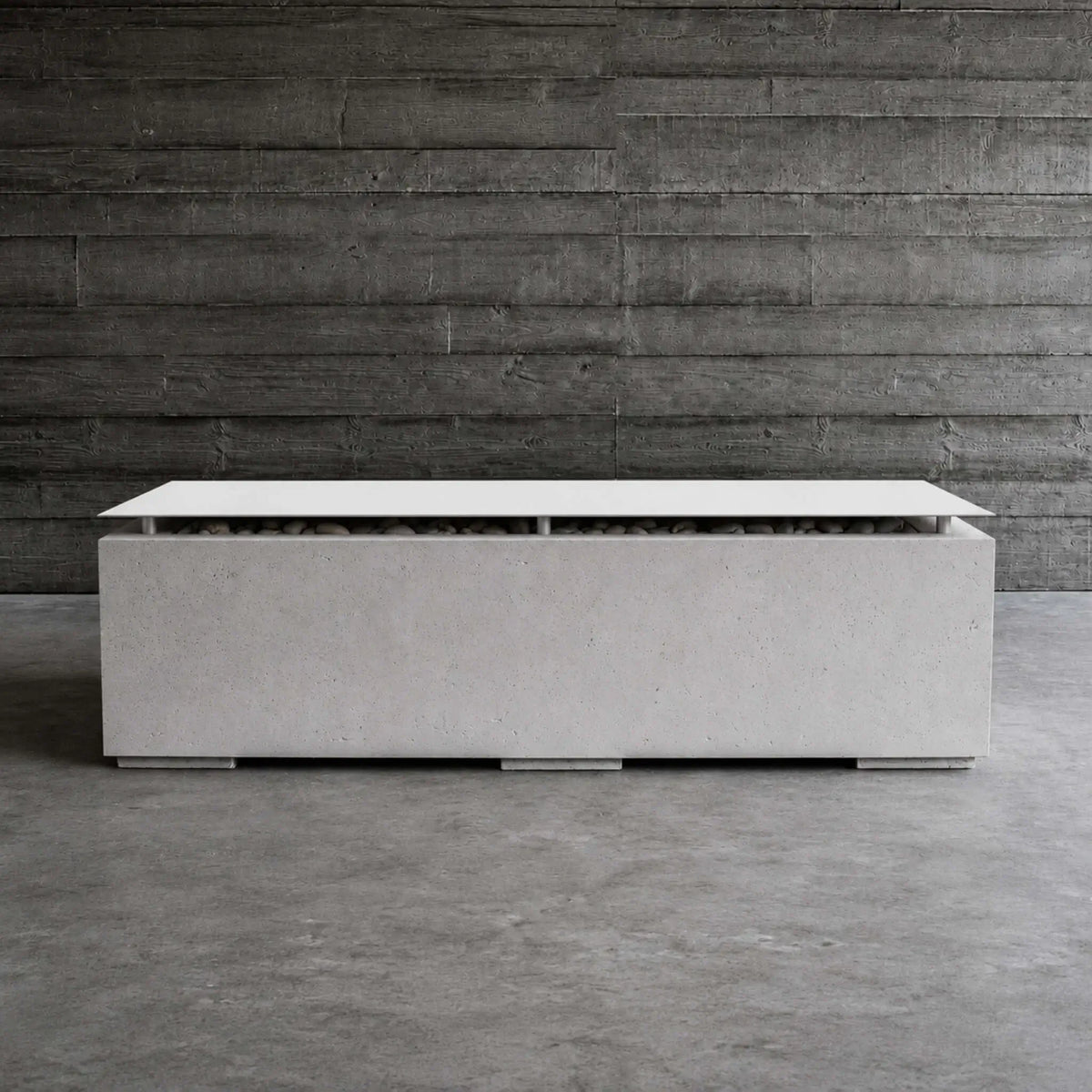 Lifestyle image of the HPC Fire Hearth Products Controls Dekko GFRC Concrete Avera 60 x 26 Fire Pit with table top lid installed. The modern rectangular fire pit sits on a concrete floor against a dark reclaimed wood wall, highlighting its light gray GFRC concrete finish, stainless steel lid, concealed fire media, and upscale contemporary outdoor design aesthetic.