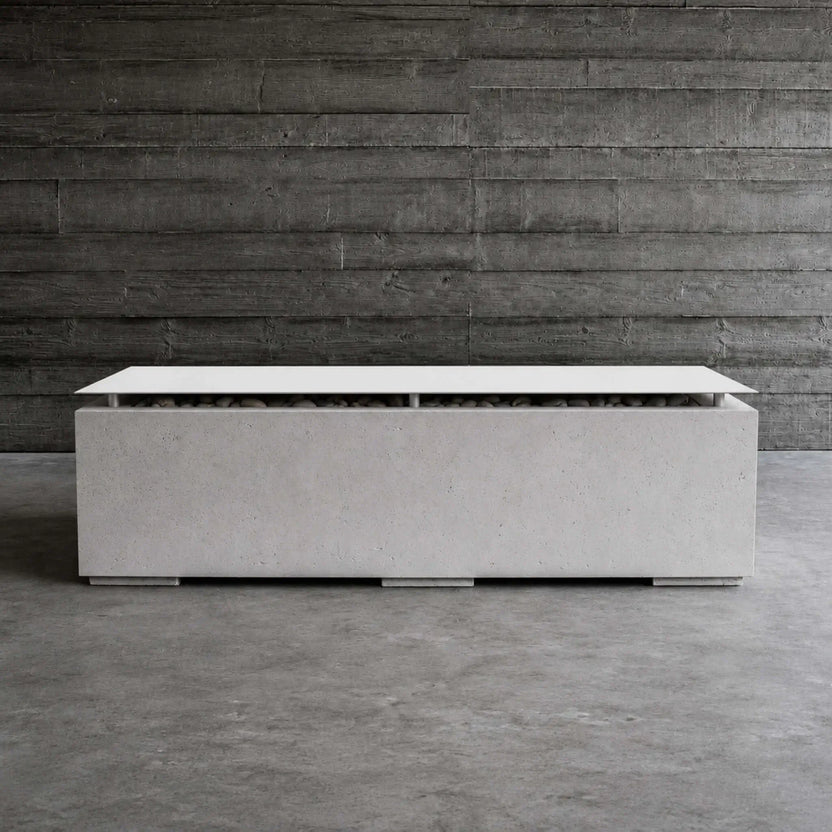 Lifestyle image of the HPC Fire Hearth Products Controls Dekko GFRC Concrete Avera 60 x 26 Fire Pit with table top lid installed. The modern rectangular fire pit sits on a concrete floor against a dark reclaimed wood wall, highlighting its light gray GFRC concrete finish, stainless steel lid, concealed fire media, and upscale contemporary outdoor design aesthetic.