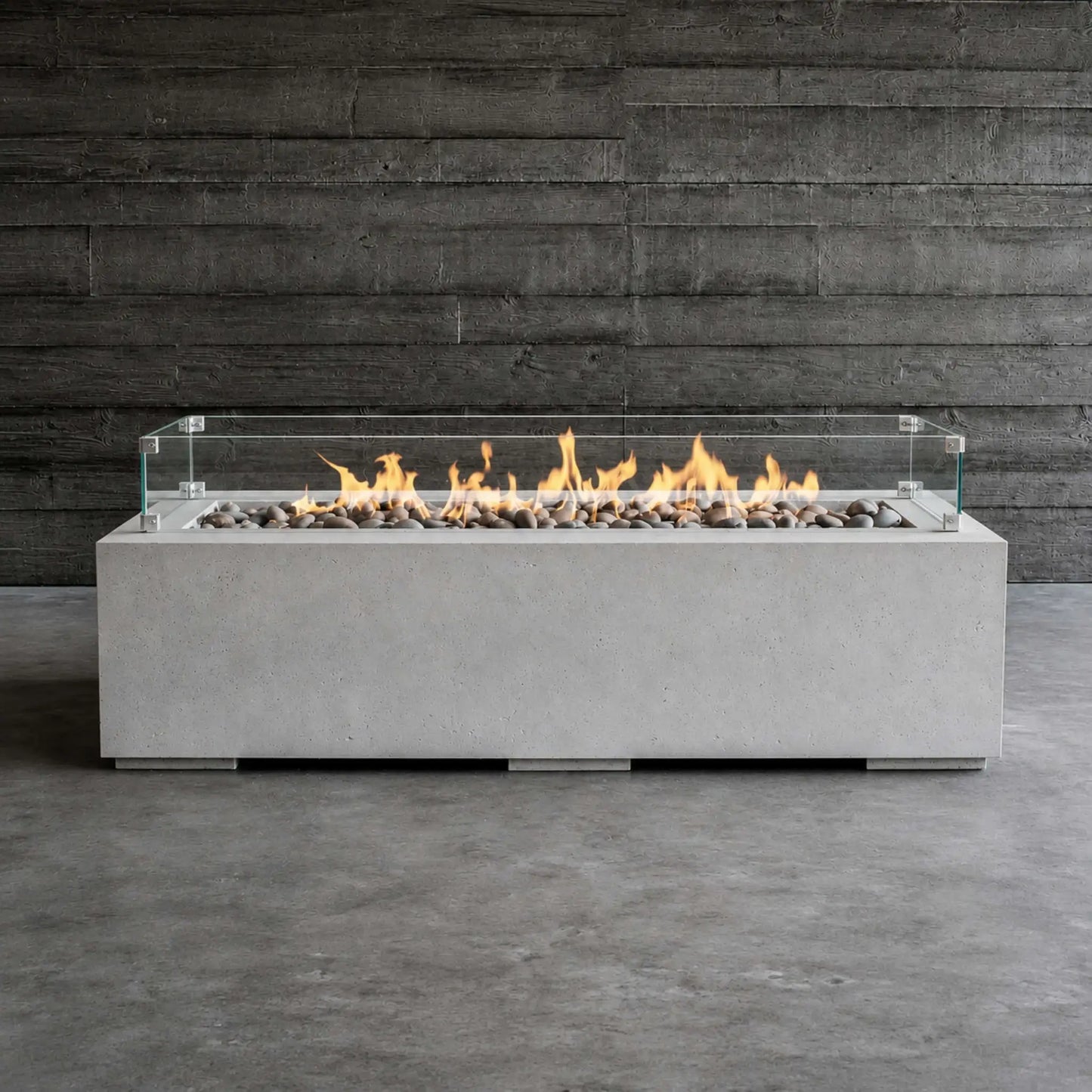 HPC Fire Hearth Products Controls Dekko GFRC Concrete Avera 60 x 26 Fire Pit Tempered Glass Wind Guard Lifestyle 1 image showing the fire pit fully lit with vibrant flames dancing above gray decorative stones. The rectangular GFRC concrete fire pit is positioned against a dark wood wall backdrop, highlighting the clear glass wind guard, modern linear burner design, and contemporary outdoor fire feature aesthetic.