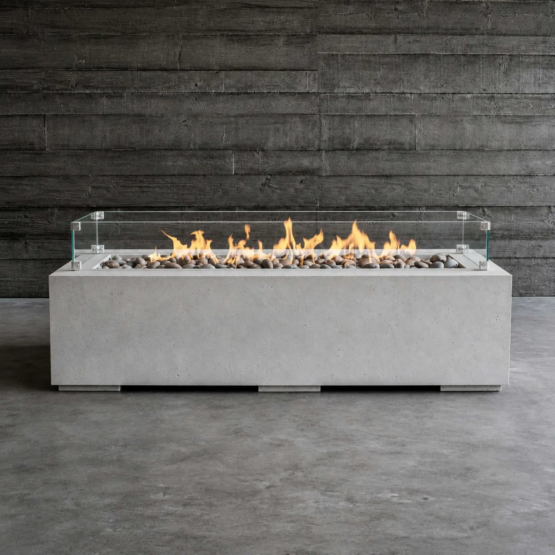 HPC Fire Hearth Products Controls Dekko GFRC Concrete Avera 60 x 26 Fire Pit Tempered Glass Wind Guard Lifestyle 1 image showing the fire pit fully lit with vibrant flames dancing above gray decorative stones. The rectangular GFRC concrete fire pit is positioned against a dark wood wall backdrop, highlighting the clear glass wind guard, modern linear burner design, and contemporary outdoor fire feature aesthetic.
