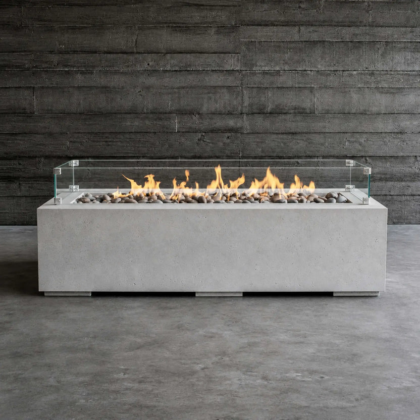 HPC Fire Hearth Products Controls Dekko GFRC Concrete Avera 60 x 26 Fire Pit Tempered Glass Wind Guard Lifestyle 1 image showing the fire pit fully lit with vibrant flames dancing above gray decorative stones. The rectangular GFRC concrete fire pit is positioned against a dark wood wall backdrop, highlighting the clear glass wind guard, modern linear burner design, and contemporary outdoor fire feature aesthetic.