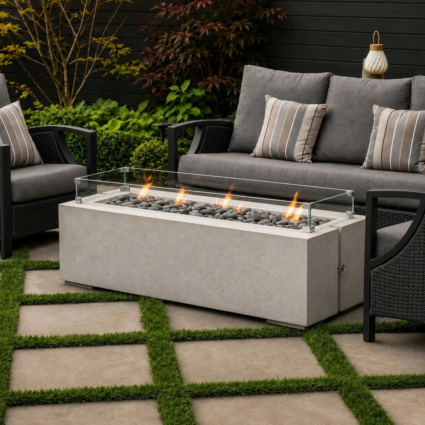 HPC Fire Hearth Products Controls Dekko GFRC Concrete Avera 60 x 26 Fire Pit Tempered Glass Wind Guard Lifestyle 2 image featuring the fire pit installed in a stylish outdoor seating area. Surrounded by gray cushioned patio furniture, landscaping, and stone pavers with grass joints, the lit linear burner and clear glass wind guard create a warm, inviting atmosphere for outdoor entertaining and backyard relaxation.