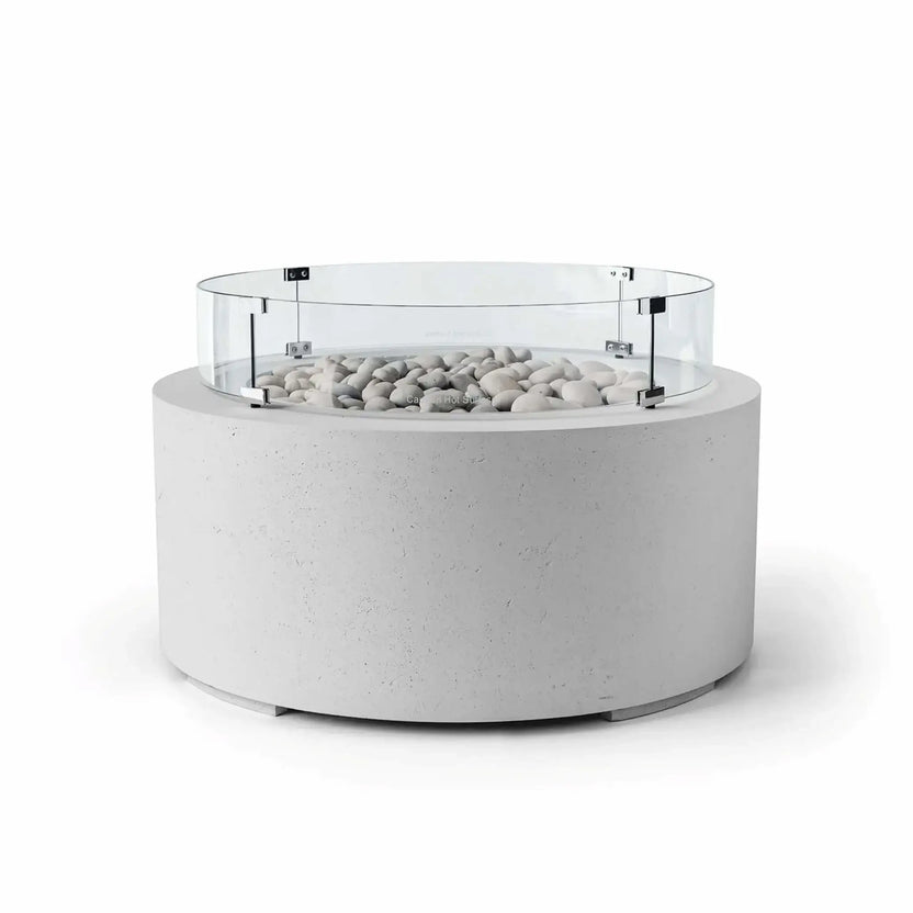 HPC Fire Hearth Products Controls Dekko GFRC Concrete Belmont Fire Pit in Ash finish featuring Ivory Stone media and a round Tempered Glass Wind Guard. The modern fire pit showcases a light gray concrete cylinder base, decorative ivory fire stones, and a clear glass windscreen secured with metal brackets. Displayed on a white background, this Belmont Fire Pit combines contemporary outdoor design, wind protection, and durable GFRC concrete construction.