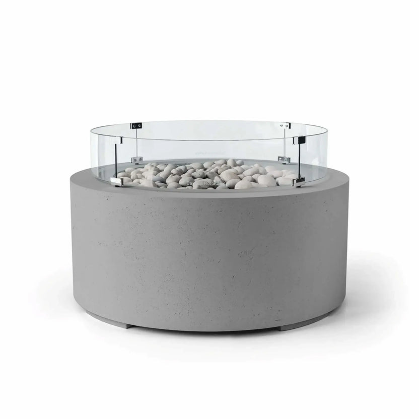 HPC Fire Hearth Products Controls Dekko GFRC Concrete Belmont Fire Pit in Charcoal finish featuring Ivory Stone media and a Tempered Glass Wind Guard. The dark gray concrete fire pit is accented by smooth ivory stones and surrounded by a clear circular glass windscreen that helps shield flames from wind. Presented on a white background, this Belmont Fire Pit offers a sleek and sophisticated centerpiece for modern outdoor living spaces.
