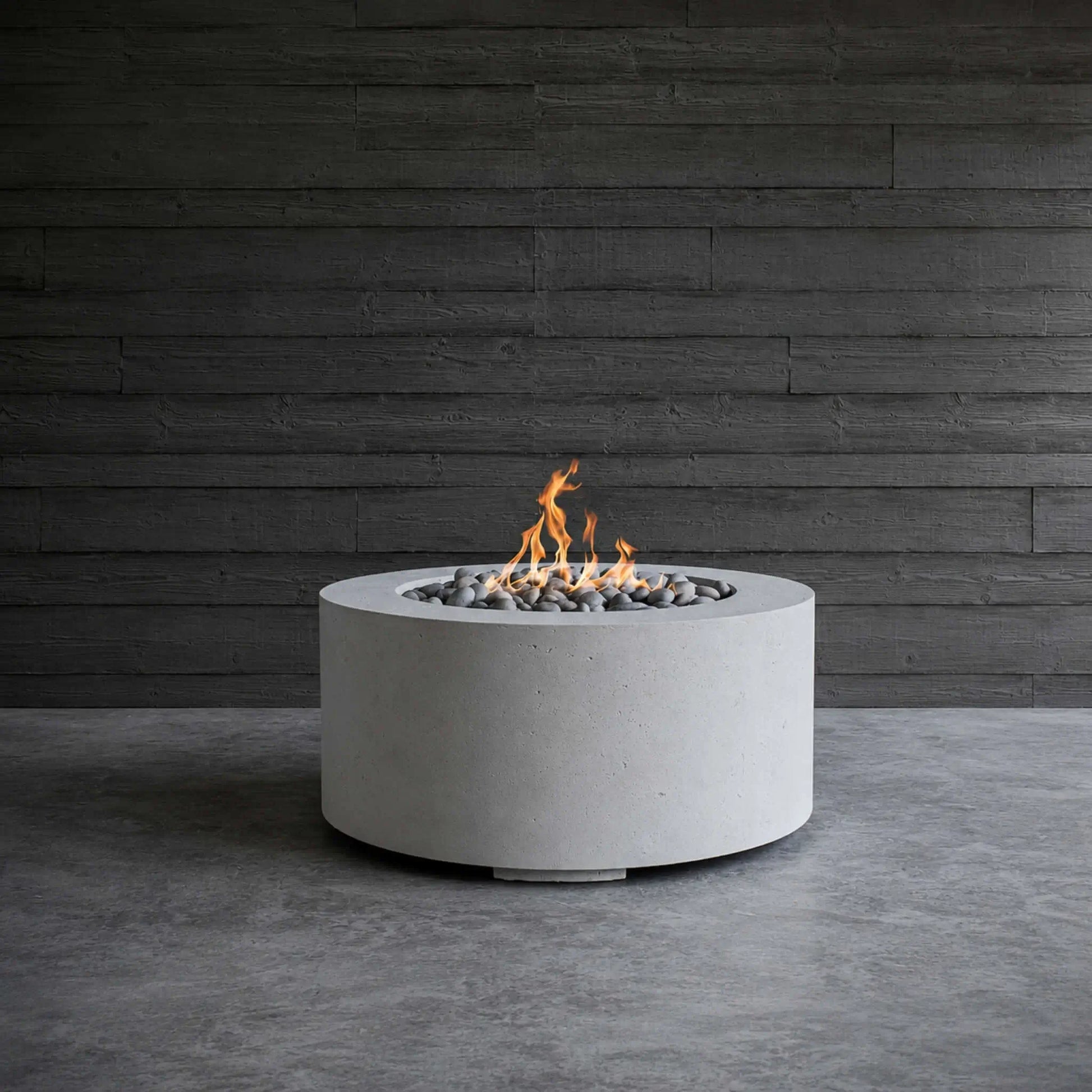 HPC Fire Hearth Products Controls Dekko GFRC Concrete Belmont Fire Pit displayed in a modern architectural setting with a dark wood accent wall backdrop. The round GFRC concrete fire pit features decorative smooth stones surrounding an active flame, highlighting the clean lines, low-profile design, and contemporary aesthetic of the Belmont Fire Pit. The image emphasizes texture, craftsmanship, and luxury outdoor fire feature styling.