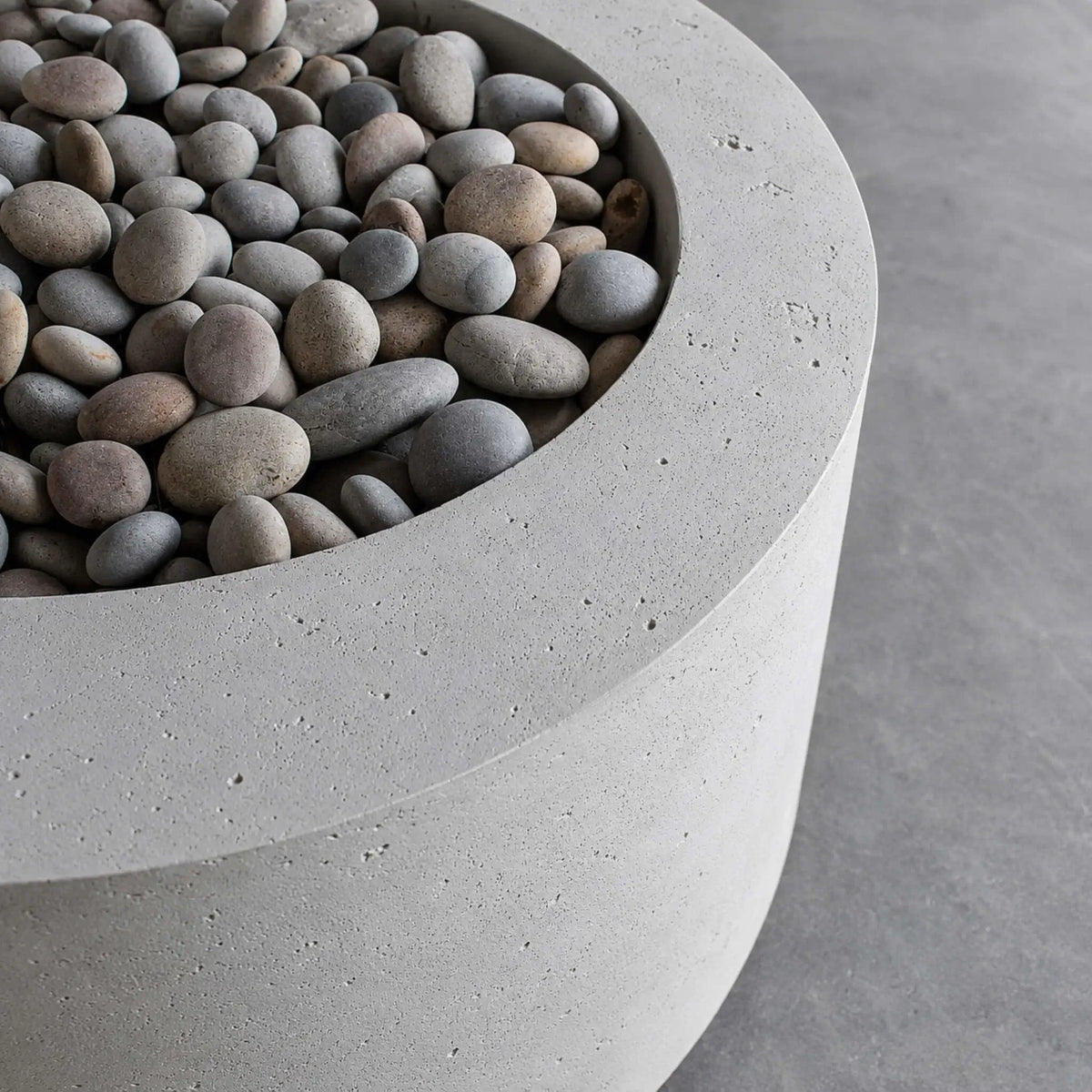 Close-up detail of the HPC Fire Hearth Products Controls Dekko GFRC Concrete Belmont Fire Pit showing the textured concrete rim and recessed fire media area filled with smooth decorative river stones. This lifestyle image highlights the premium GFRC concrete construction, subtle surface texture, rounded edges, and refined craftsmanship that define the Belmont Fire Pit’s modern outdoor design.