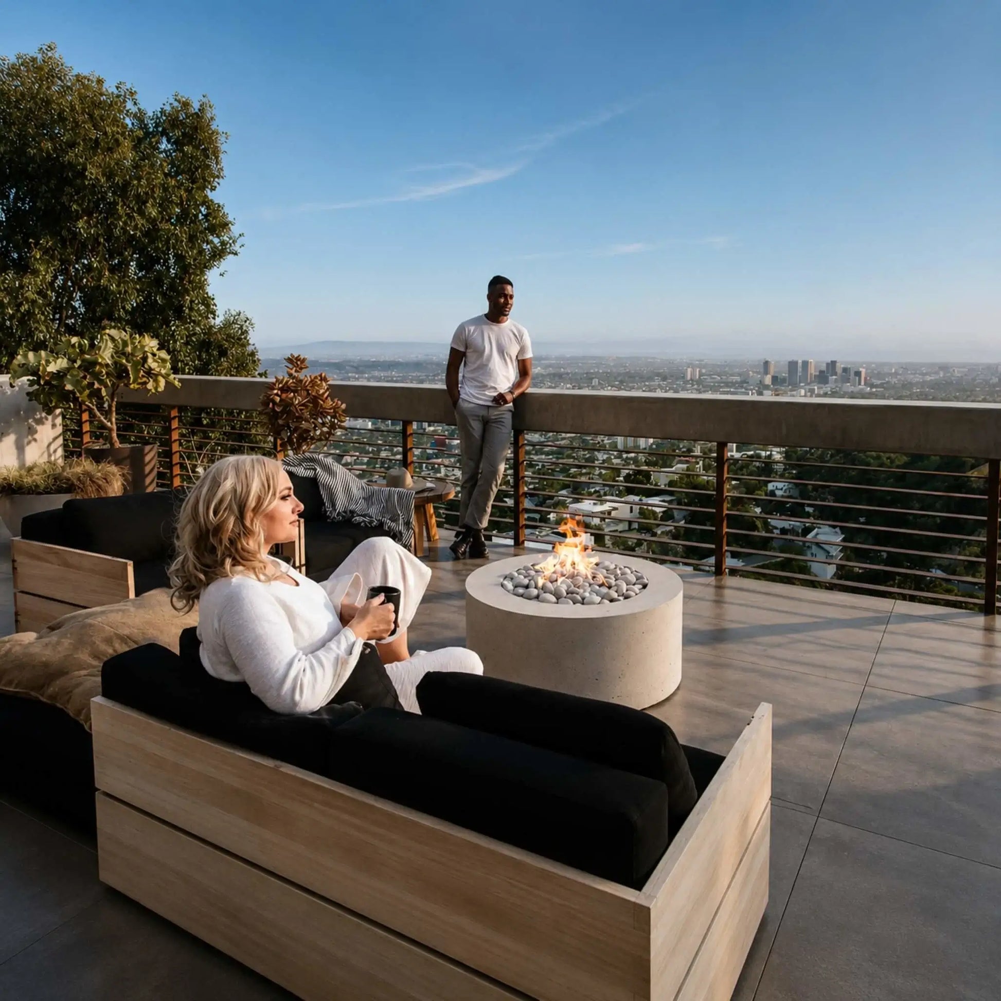 HPC Fire Hearth Products Controls Dekko GFRC Concrete Belmont Fire Pit installed on a luxury rooftop terrace overlooking a city skyline. The round fire pit glows with an active flame surrounded by smooth stones while guests relax on modern outdoor furniture. The Belmont Fire Pit serves as a contemporary centerpiece, enhancing the rooftop outdoor living space with warmth, ambiance, and sophisticated design.
