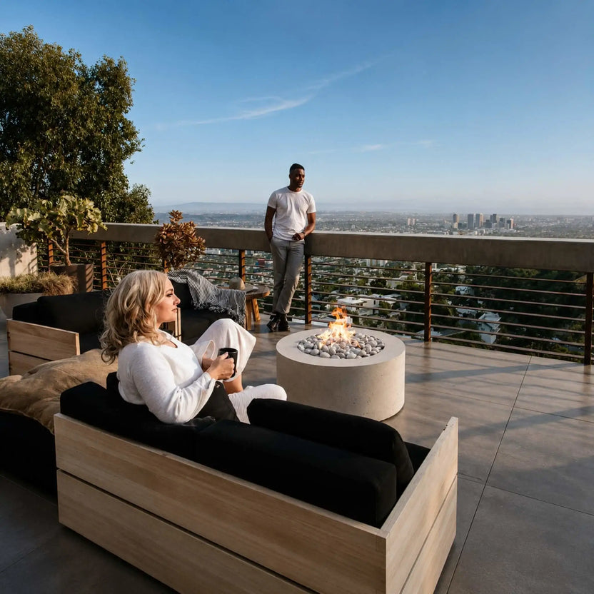 HPC Fire Hearth Products Controls Dekko GFRC Concrete Belmont Fire Pit installed on a luxury rooftop terrace overlooking a city skyline. The round fire pit glows with an active flame surrounded by smooth stones while guests relax on modern outdoor furniture. The Belmont Fire Pit serves as a contemporary centerpiece, enhancing the rooftop outdoor living space with warmth, ambiance, and sophisticated design.