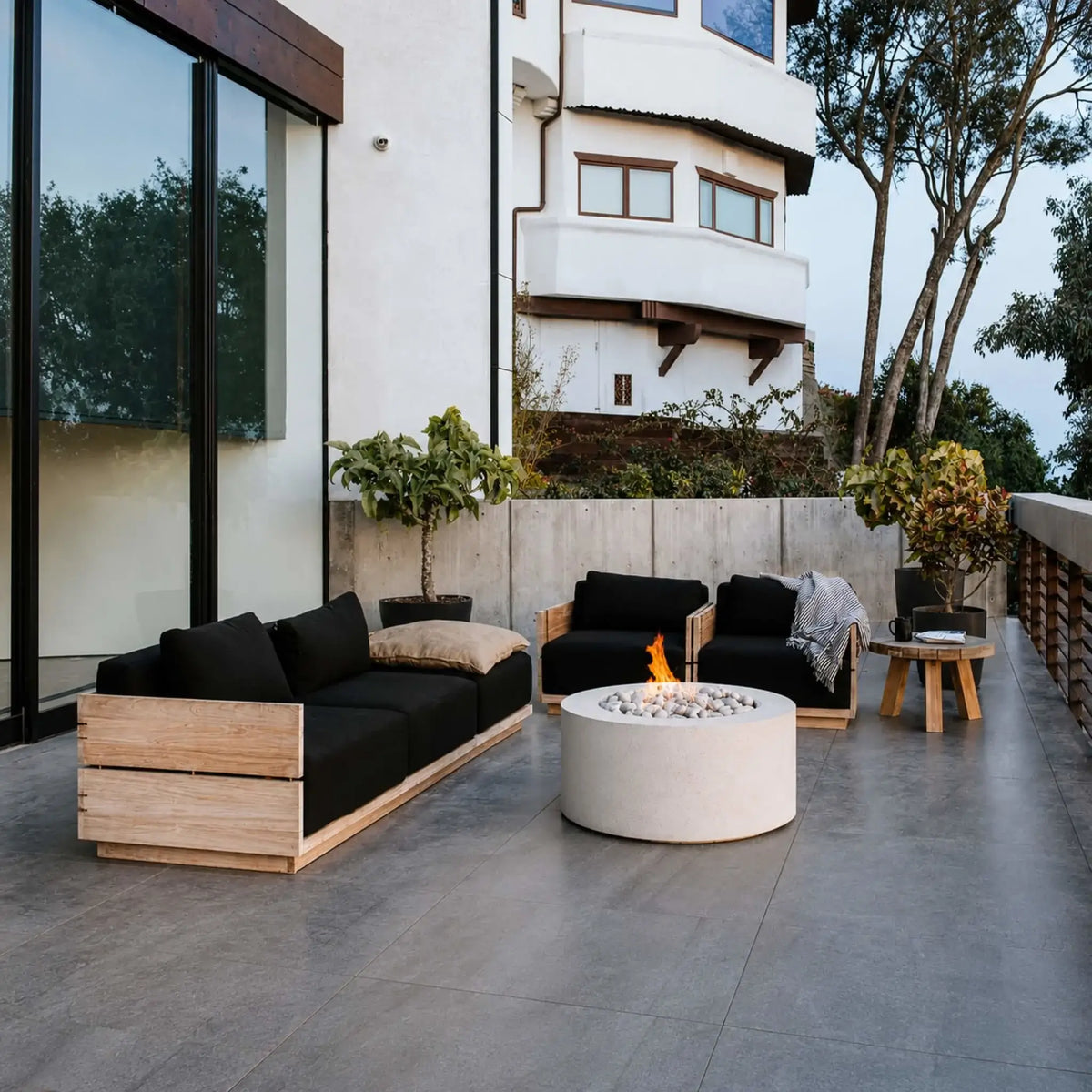 HPC Fire Hearth Products Controls Dekko GFRC Concrete Belmont Fire Pit positioned in the center of a contemporary outdoor patio furnished with wood-framed seating and black cushions. The round concrete fire pit features a natural flame rising above decorative stone media, creating a welcoming gathering space. Modern architecture, clean lines, and landscaped surroundings highlight the Belmont Fire Pit’s upscale outdoor appeal.