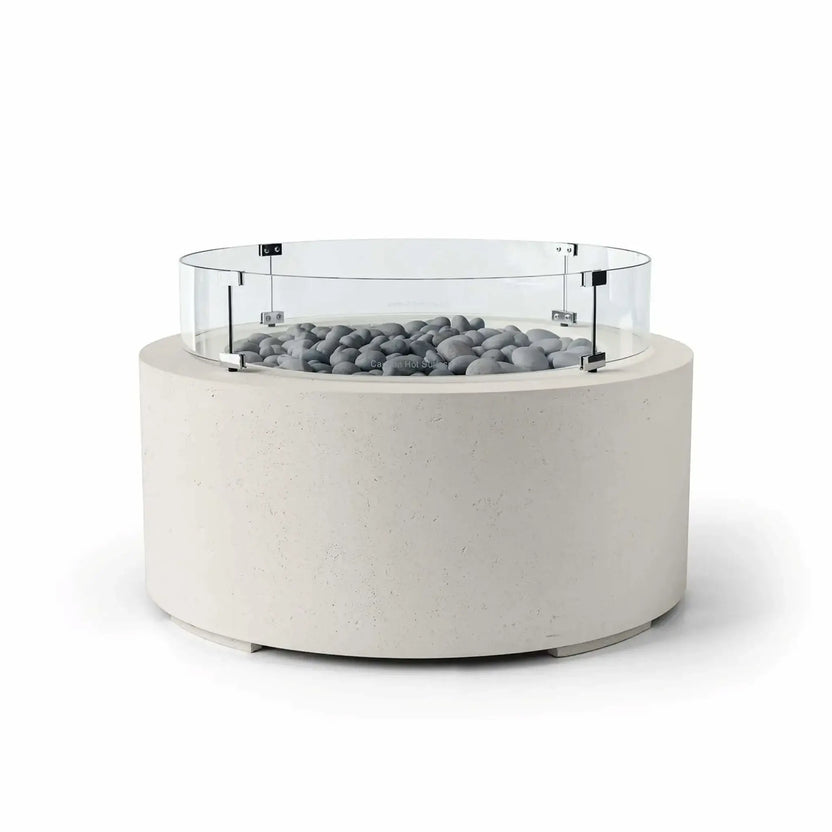 HPC Fire Hearth Products Controls Dekko GFRC Concrete Belmont Fire Pit in Limestone finish featuring Slate Stone fire media and a Tempered Glass Wind Guard. This contemporary round fire pit showcases a light limestone-colored concrete body, smooth gray slate stones around the burner area, and a transparent circular glass windscreen supported by metal hardware. The minimalist design makes the Belmont Fire Pit an elegant addition to modern outdoor entertaining areas.