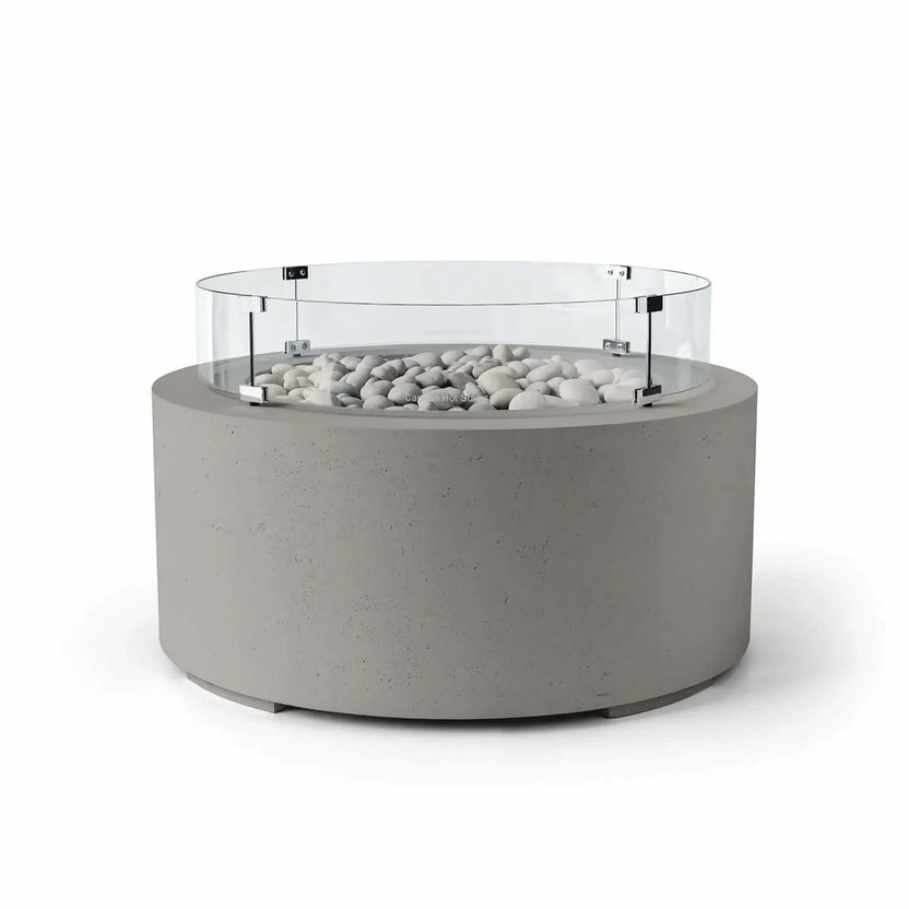 HPC Fire Hearth Products Controls Dekko GFRC Concrete Belmont Fire Pit in Sandstone finish with Ivory Stone fire media and a Tempered Glass Wind Guard. The warm beige sandstone-colored concrete base is paired with smooth ivory stones and surrounded by a circular clear glass windscreen that helps shield flames from wind. Displayed on a white background, this Belmont Fire Pit brings natural warmth and modern design to outdoor living environments.