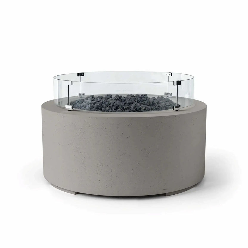 HPC Fire Hearth Products Controls Dekko GFRC Concrete Belmont Fire Pit in Sandstone finish featuring Lava Rock media and a Tempered Glass Wind Guard. The round fire pit showcases a textured sandstone-colored GFRC concrete exterior, dark volcanic lava rock fire media, and a transparent circular windscreen mounted with stainless steel brackets. The Belmont Fire Pit offers a sophisticated blend of natural textures and contemporary outdoor styling.