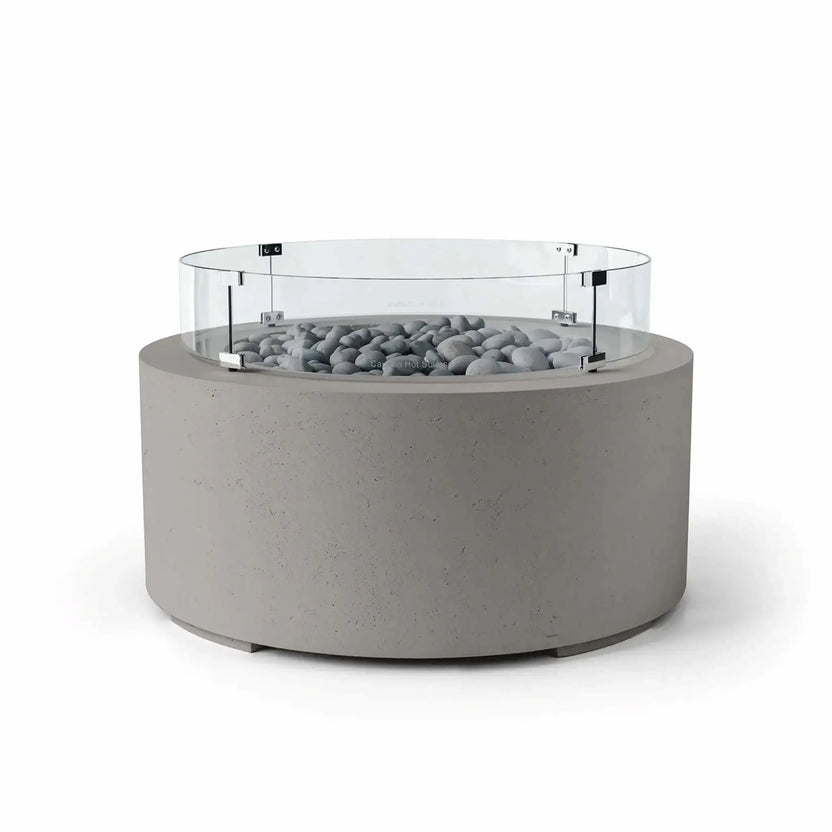 HPC Fire Hearth Products Controls Dekko GFRC Concrete Belmont Fire Pit in Sandstone finish with Slate Stone fire media and a Tempered Glass Wind Guard. This modern round fire pit features a warm beige concrete body, smooth gray slate stones surrounding the burner area, and a clear circular glass windscreen designed for enhanced flame protection. The Belmont Fire Pit serves as an elegant outdoor centerpiece for patios, terraces, and lounge areas.
