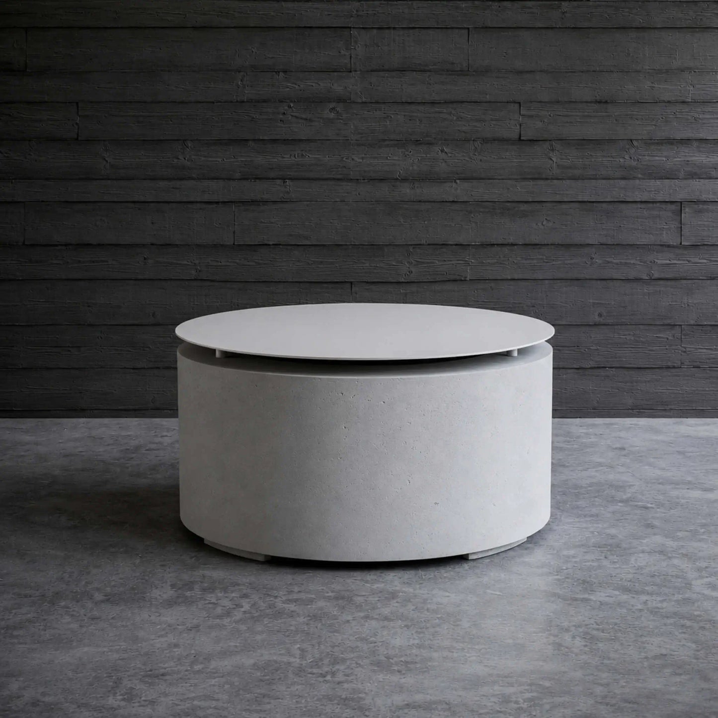 HPC Fire Hearth Products Controls Dekko GFRC Concrete Belmont Fire Pit Table Top Lid displayed in a modern outdoor lounge setting. The round concrete fire pit with lid serves as a stylish coffee table surrounded by contemporary wood-framed seating with black cushions. Positioned on a spacious patio with architectural landscaping and modern home design elements, the Belmont Fire Pit demonstrates its versatility as both a fire feature and functional outdoor furniture piece.