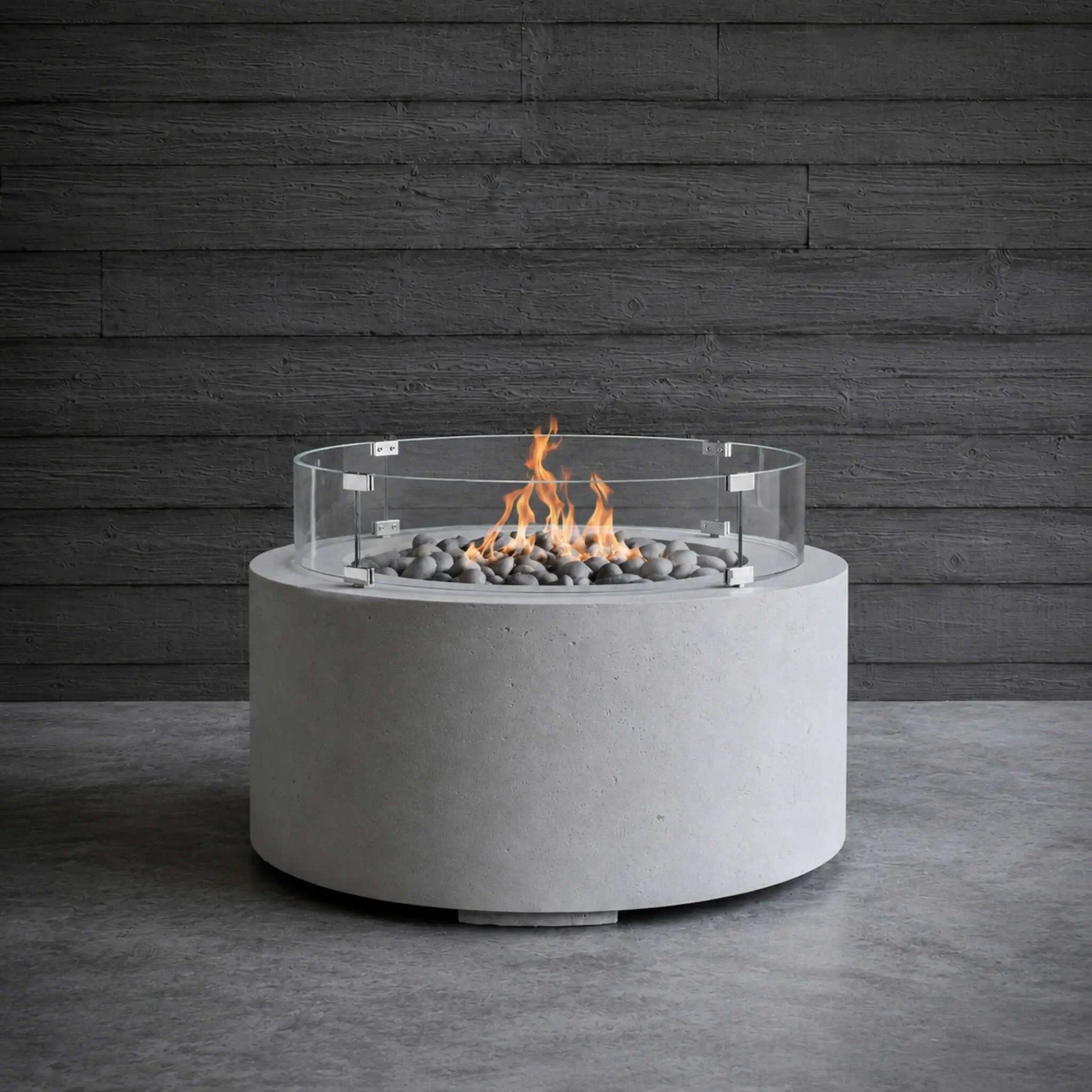 HPC Fire Hearth Products Controls Dekko GFRC Concrete Belmont Fire Pit with Tempered Glass Wind Guard shown in a lifestyle setting. The round concrete fire pit features gray stone media and active flames protected by a clear glass windscreen. Positioned on a concrete floor against a dark wood plank wall, the Belmont Fire Pit highlights its contemporary design, durable GFRC concrete construction, and inviting ambiance for modern outdoor entertaining spaces.