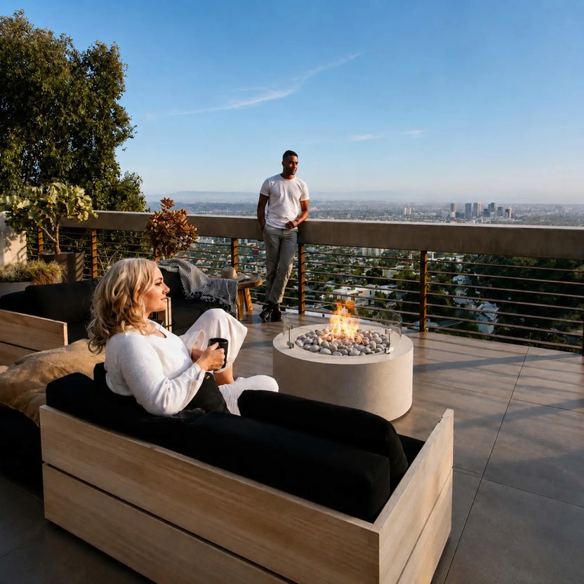 HPC Fire Hearth Products Controls Dekko GFRC Concrete Belmont Fire Pit with Tempered Glass Wind Guard featured on a luxury rooftop patio overlooking a city skyline. A woman relaxes on outdoor seating while a man stands near the railing, enjoying the warmth of the Belmont Fire Pit. The round concrete fire feature includes decorative stone media, a clear glass windscreen, and bright flames, creating a stylish focal point for upscale outdoor living and entertaining.