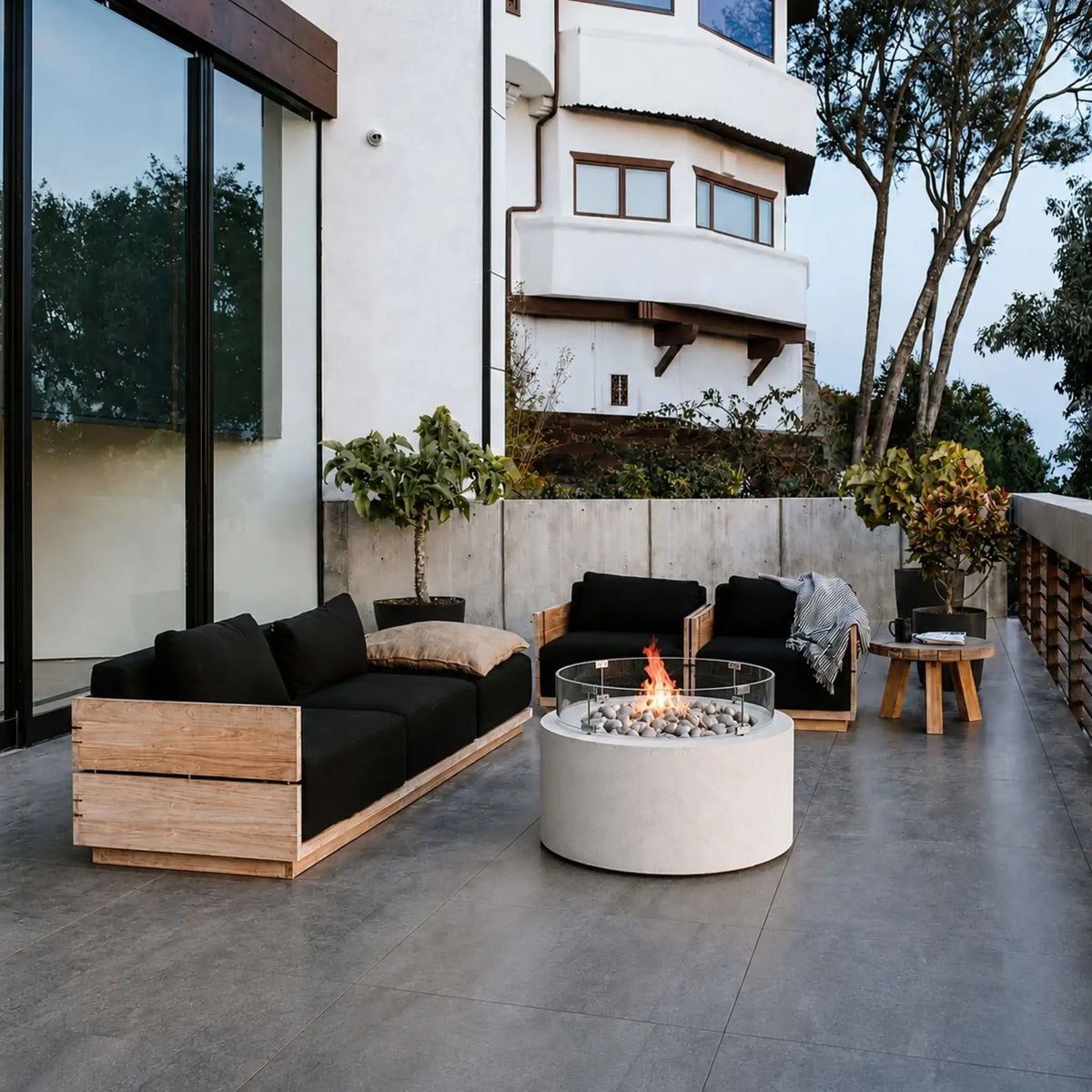 HPC Fire Hearth Products Controls Dekko GFRC Concrete Belmont Fire Pit with Tempered Glass Wind Guard showcased in a modern outdoor patio lifestyle setting. The round Belmont Fire Pit features a smooth white GFRC concrete base, decorative gray fire stones, and a clear circular glass windscreen surrounding a lively flame. Positioned between contemporary wood-framed lounge seating with black cushions, the fire pit creates a warm focal point on a spacious tiled terrace beside a luxury home.