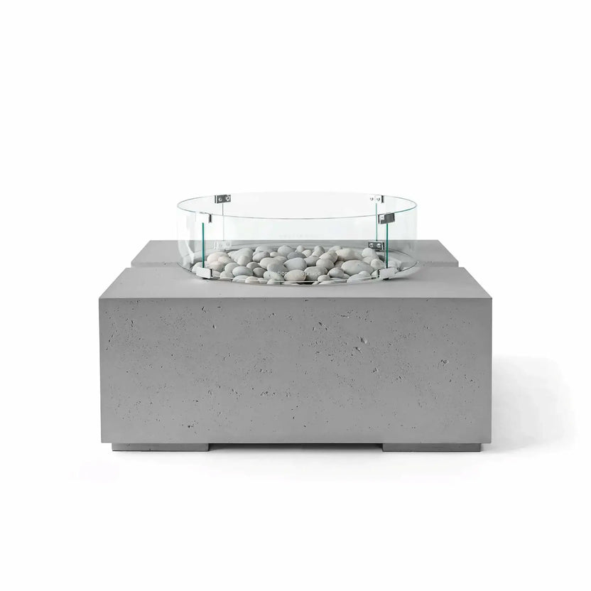 HPC Fire Hearth Products Controls Dekko GFRC Concrete Bravo 32 x 32 Fire Pit in Charcoal finish with Ivory Stone fire media and a tempered glass wind guard. The dark gray textured concrete body contrasts beautifully with the light decorative stones, highlighting the fire pit’s modern geometric design and premium outdoor living appeal in a studio-style product image.