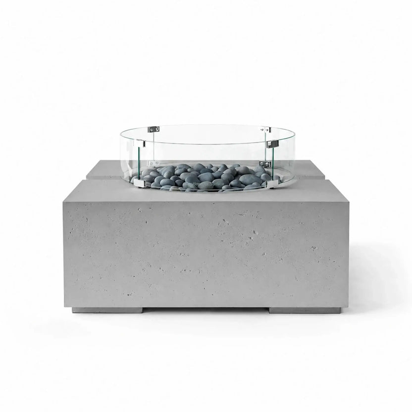 HPC Fire Hearth Products Controls Dekko GFRC Concrete Bravo 32 x 32 Fire Pit in Charcoal finish with Slate Stone fire media and a tempered glass wind guard. The dark concrete base and smooth gray stones create a sleek contemporary look, while the circular glass guard surrounds the burner area, emphasizing safety and modern outdoor design.