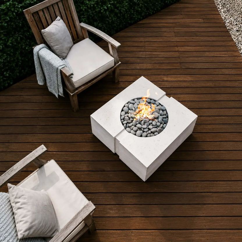 Overhead lifestyle view of the HPC Fire Hearth Products Controls Dekko GFRC Concrete Bravo 32 x 32 Fire Pit installed on a rich wood deck. The modern square concrete fire pit features smooth gray slate stones surrounding an active flame and is paired with cushioned wooden lounge chairs, creating a warm and inviting outdoor living space with contemporary design appeal.