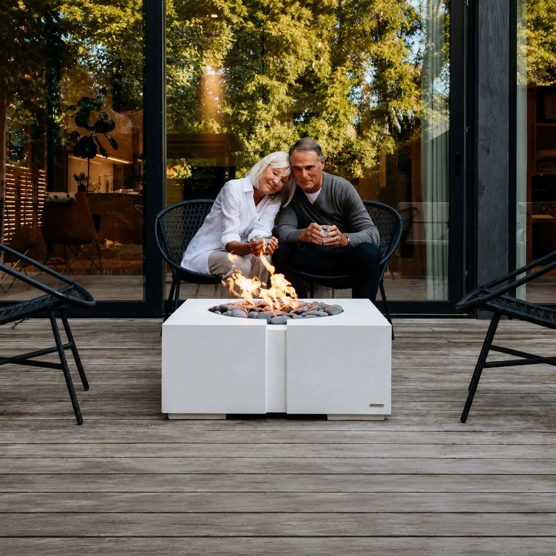Lifestyle image of the Dekko GFRC Concrete Bravo 32 x 32 Fire Pit by HPC Fire Hearth Products Controls positioned on a modern residential deck. A mature couple relaxes beside the fire feature, which showcases a square concrete design, decorative gray fire stones, and vibrant flames. Large glass walls and surrounding trees create a comfortable outdoor gathering environment.