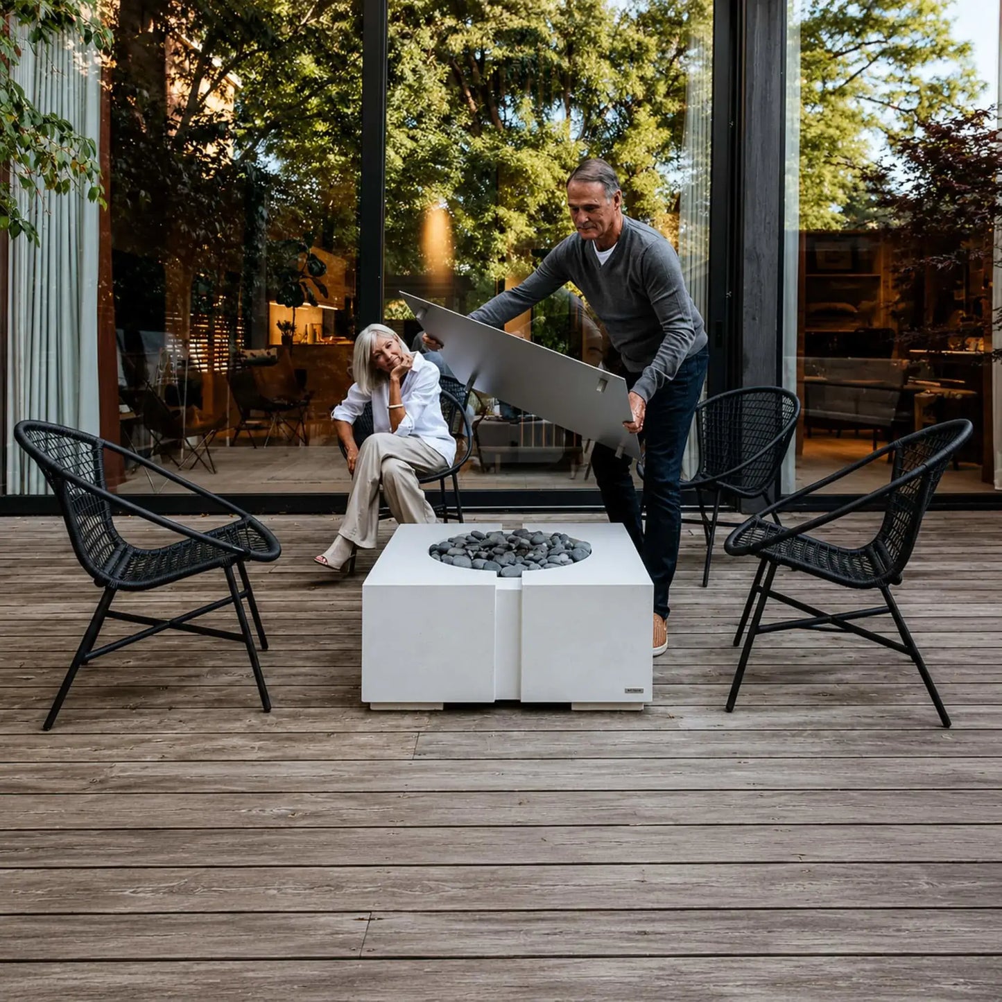 HPC Dekko GFRC Concrete Bravo 32 x 32 Fire Pit shown in a real-world outdoor setting with a removable tabletop cover being lifted to reveal the recessed fire media area. The image highlights the fire pit’s dual-purpose functionality as both a fire feature and outdoor table, surrounded by modern patio seating on a spacious wood deck.