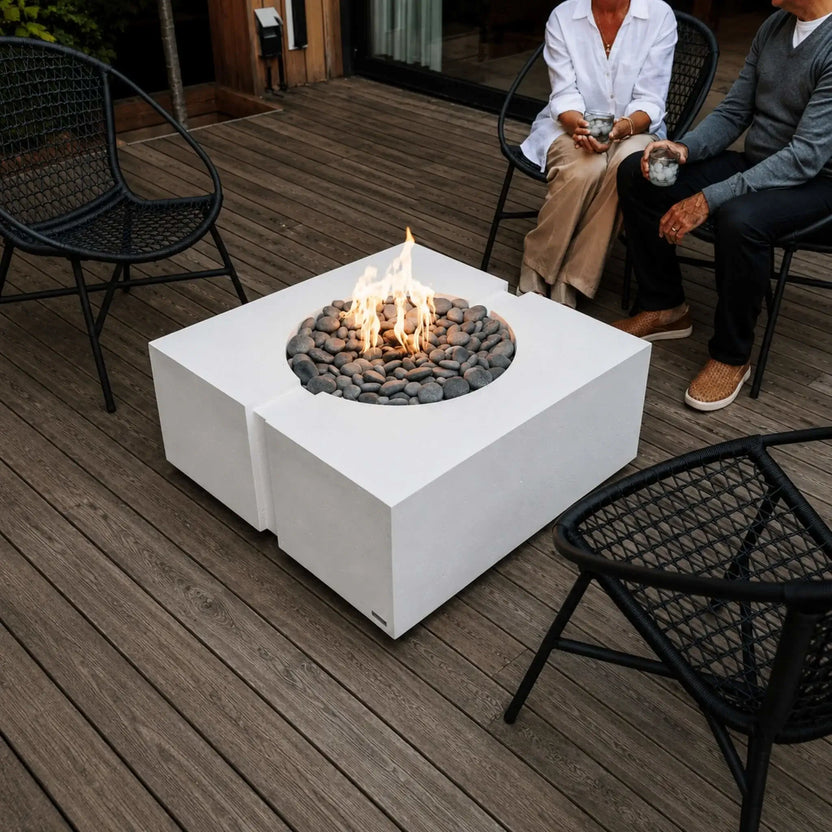 Close-up lifestyle scene featuring the HPC Fire Hearth Products Controls Dekko GFRC Concrete Bravo 32 x 32 Fire Pit with active flames rising through decorative gray stones. The square concrete fire pit is surrounded by black woven patio chairs and seated guests enjoying drinks, showcasing a cozy outdoor entertainment area on a natural wood deck.