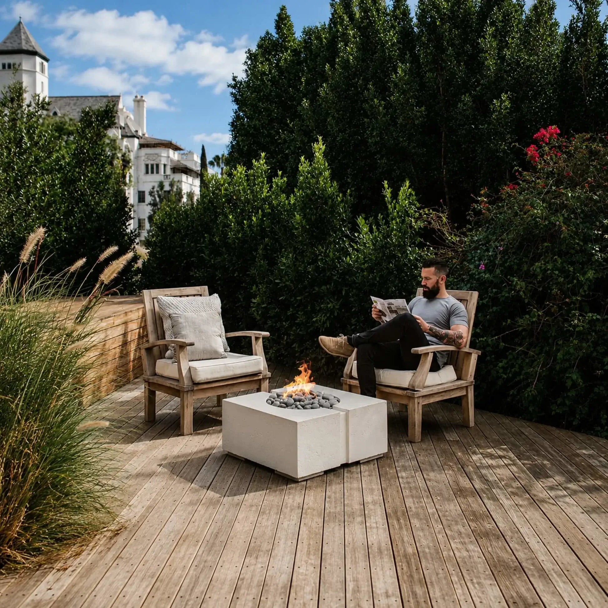 Outdoor lifestyle photograph of the Dekko GFRC Concrete Bravo 32 x 32 Fire Pit from HPC Fire Hearth Products Controls placed between two cushioned wooden lounge chairs. A man relaxes while reading beside the lit fire pit, surrounded by lush landscaping and privacy hedges, demonstrating the fire feature’s role as a stylish focal point in a backyard retreat.