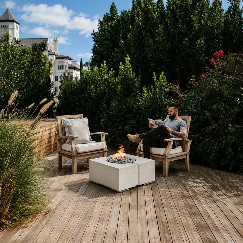 Outdoor lifestyle photograph of the Dekko GFRC Concrete Bravo 32 x 32 Fire Pit from HPC Fire Hearth Products Controls placed between two cushioned wooden lounge chairs. A man relaxes while reading beside the lit fire pit, surrounded by lush landscaping and privacy hedges, demonstrating the fire feature’s role as a stylish focal point in a backyard retreat.