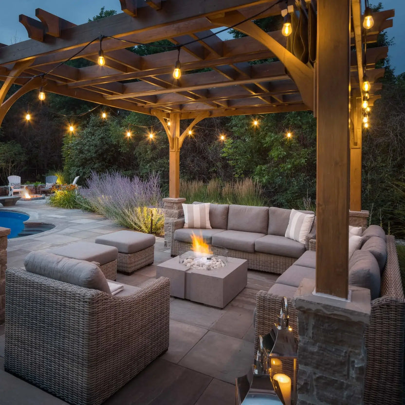 Evening outdoor living space featuring the HPC Fire Hearth Products Controls Dekko GFRC Concrete Bravo 32 x 32 Fire Pit beneath a timber pergola with hanging string lights. The illuminated fire pit sits among wicker patio furniture and cushions beside a pool, creating a warm, upscale backyard gathering area designed for relaxation and entertaining.