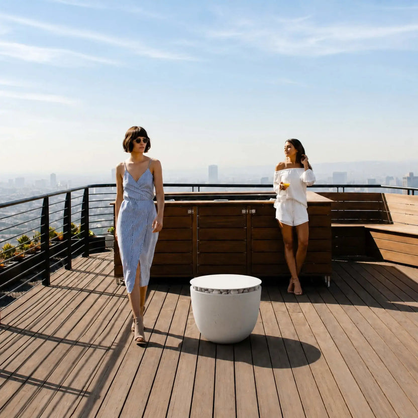 HPC Fire Hearth Products Controls Dekko GFRC Concrete Element 20 Round Fire Column Table Top Lid Lifestyle image showing the covered fire column on a luxury rooftop deck overlooking a city skyline. Two women relax near an outdoor kitchen while the round concrete fire feature is converted into a functional table using the tabletop lid. The scene demonstrates how the accessory enhances usability while maintaining a sophisticated outdoor design.