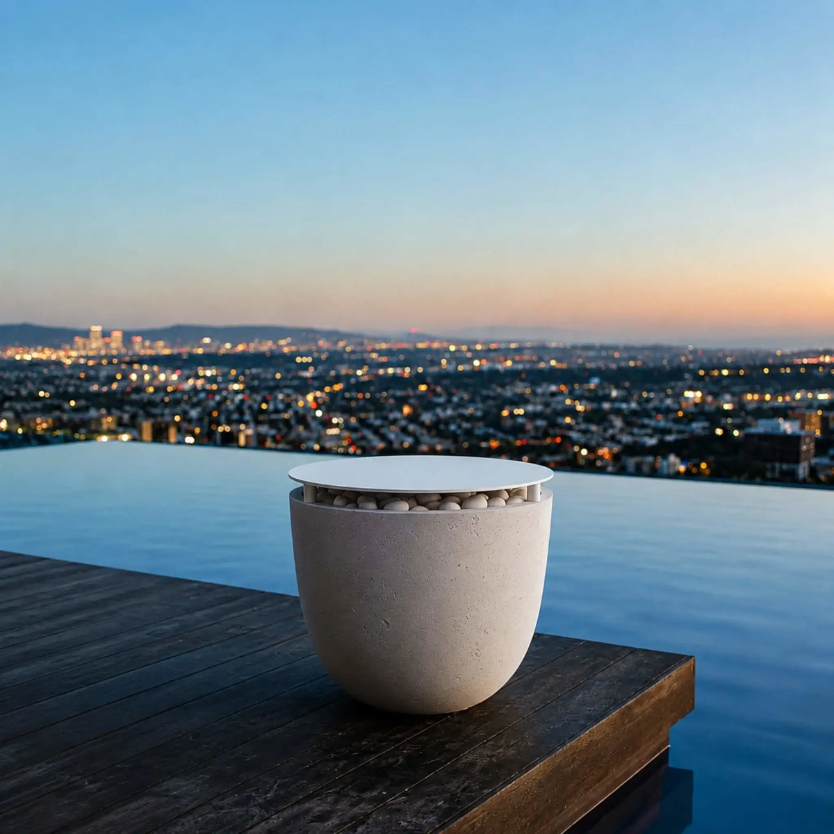 Dekko GFRC Concrete Element 20 Round Fire Column Table Top Lid Lifestyle scene from HPC Fire Hearth Products Controls featuring the covered fire column beside an infinity-edge pool at sunset. The round concrete unit is transformed into a practical outdoor table with the tabletop lid installed, complementing the luxurious poolside setting and panoramic city views. The image showcases the product’s versatility in upscale outdoor living environments.