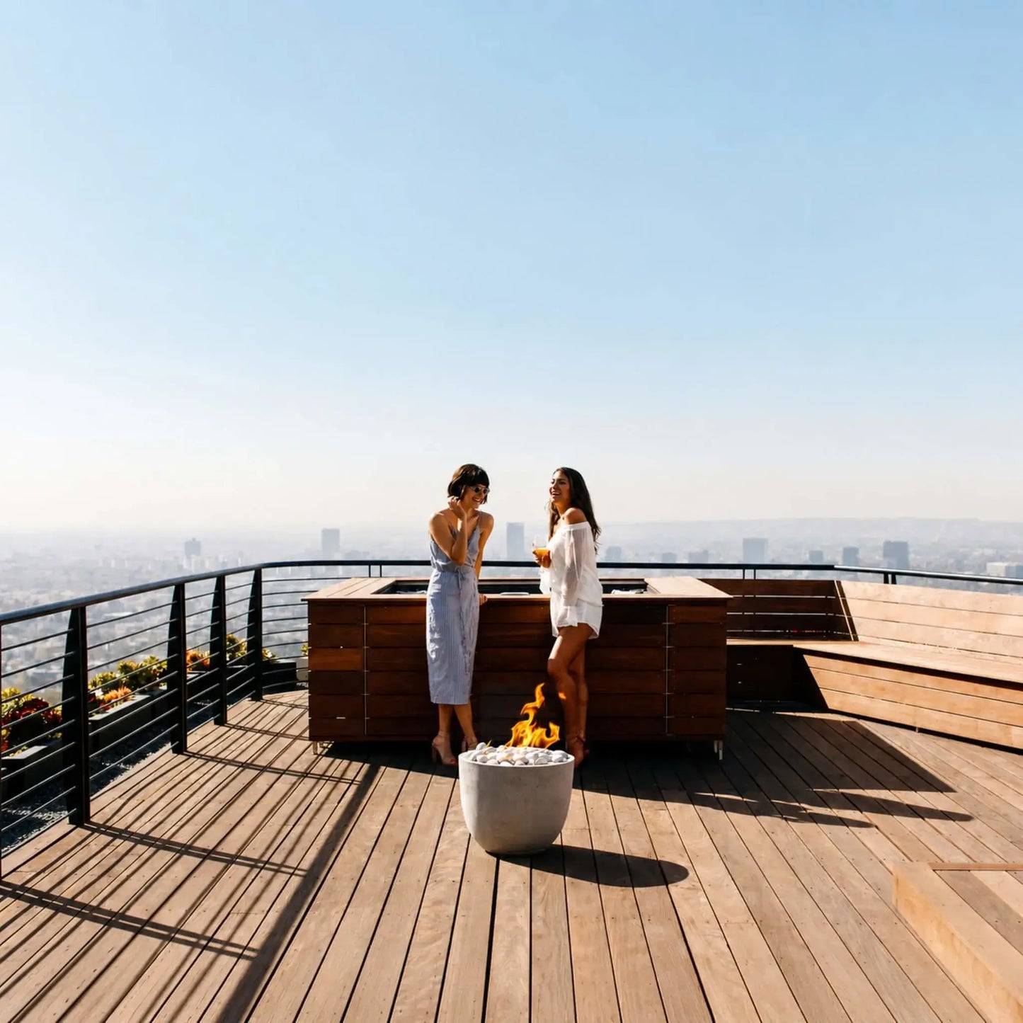 HPC Fire Hearth Products Controls Dekko GFRC Concrete Element Round 20 Fire Column showcased on a luxury rooftop deck overlooking a city skyline. Two women stand near an outdoor kitchen and seating area while a compact round GFRC concrete fire column burns with bright flames above smooth decorative stones. The modern fire feature serves as a stylish focal point for elevated outdoor entertaining spaces with panoramic urban views.