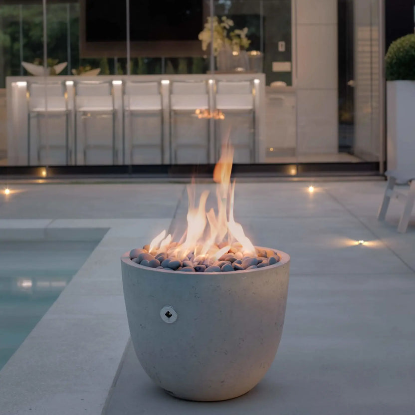 Dekko GFRC Concrete Element Round 20 Fire Column by HPC Fire Hearth Products Controls featured beside a modern pool and outdoor bar area at dusk. The rounded concrete fire feature is filled with decorative stones and produces tall natural flames that illuminate the surrounding patio. Contemporary architecture, bar seating, landscape lighting, and poolside ambiance showcase the fire column’s role in luxury outdoor living design.