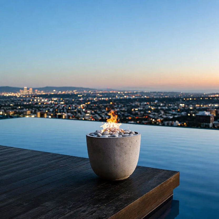HPC Dekko GFRC Concrete Element Round 20 Fire Column positioned beside an infinity-edge pool overlooking a sprawling cityscape at dusk. The round concrete fire feature is topped with smooth white stones and a vibrant flame that reflects against the water. This luxury outdoor setting showcases the fire column as an elegant accent for modern poolside patios, rooftop decks, and upscale entertaining environments.