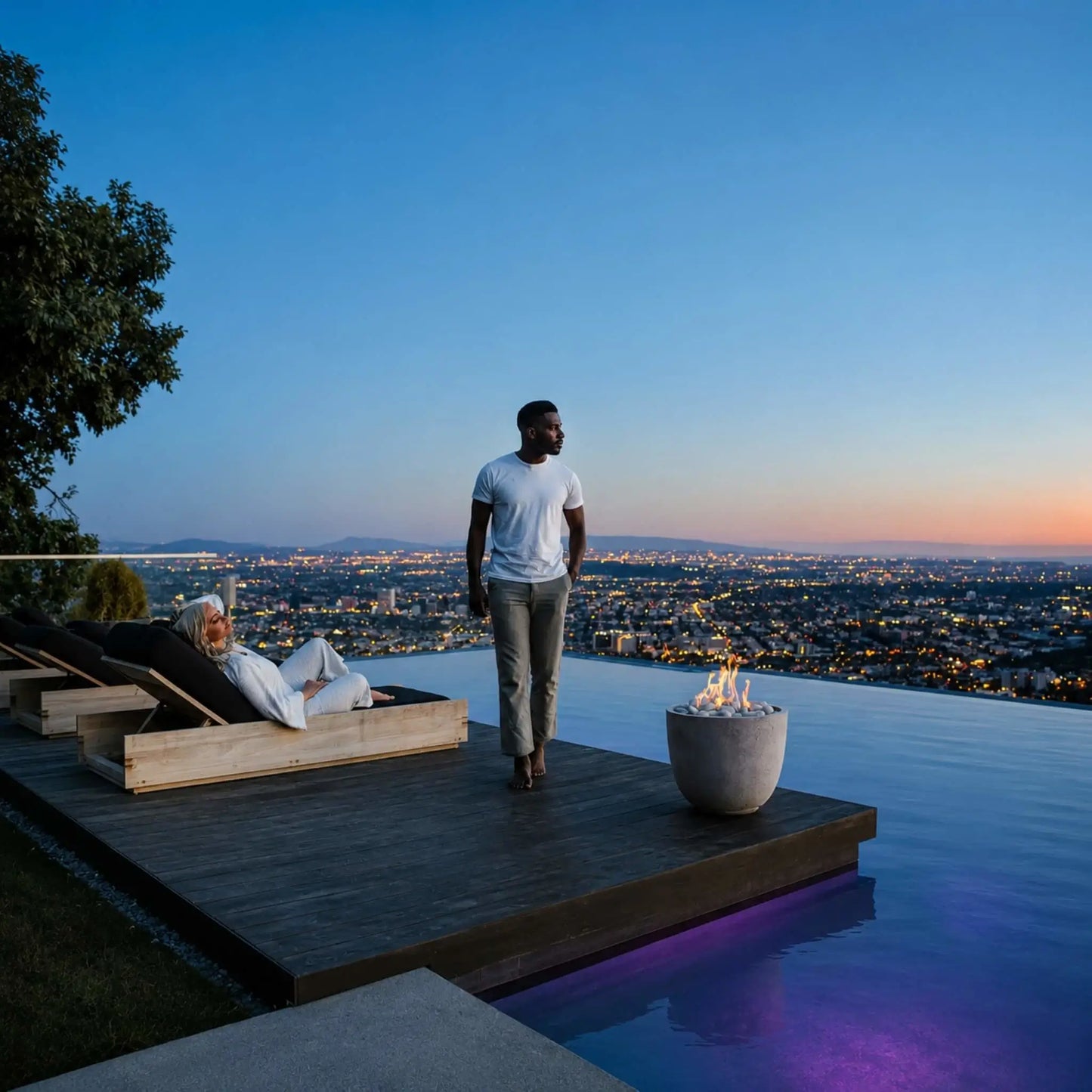 Dekko GFRC Concrete Element Round 20 Fire Column from HPC Fire Hearth Products Controls featured on a luxury infinity pool deck at sunset. A couple relaxes while enjoying panoramic city views as the compact concrete fire column glows with natural flames above decorative stone media. The image highlights the fire feature’s ability to create warmth, ambiance, and visual appeal in premium outdoor living spaces.
