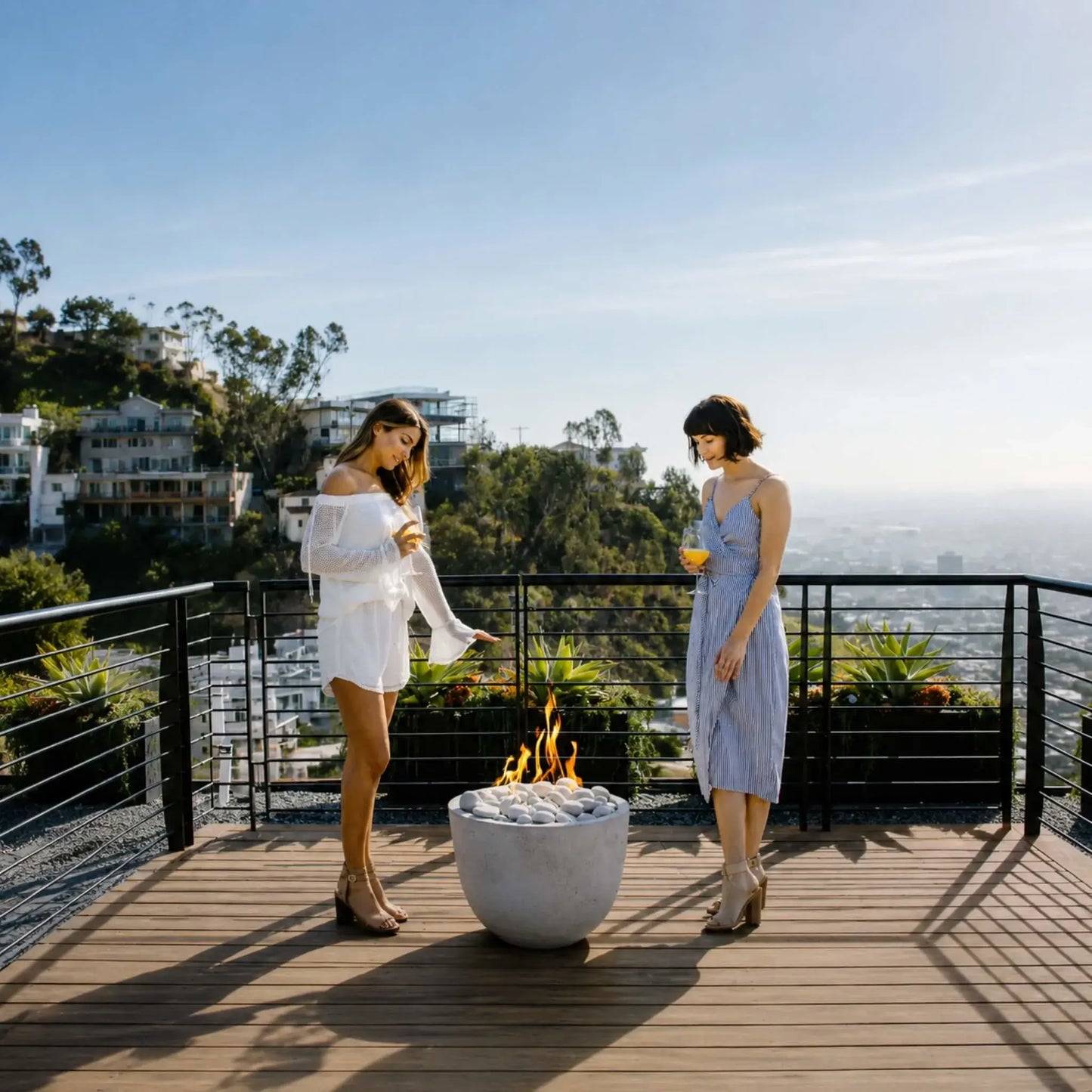 HPC Dekko GFRC Concrete Element Round 20 Fire Column serving as the centerpiece of a contemporary rooftop gathering space. A sleek round concrete fire feature with white decorative stones produces a vibrant flame while guests relax nearby against a panoramic skyline backdrop. The image emphasizes the fire column’s compact footprint, modern styling, and ability to enhance upscale outdoor living environments.